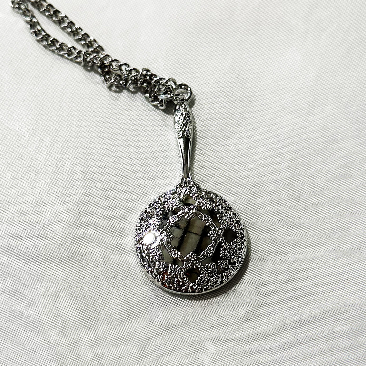 The PVRIS silver pendant necklace features a circular, ornate filigree design with a raised intricate pattern and shiny finish, displayed on a white textured surface.